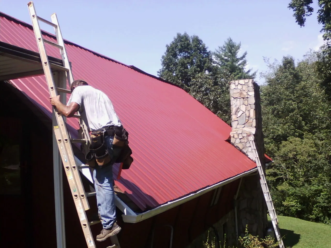 Roofer on a ladder beside a red metal roof and stone chimney — Soffit & Fascia Repair by Roofing Brothers in Greentown
